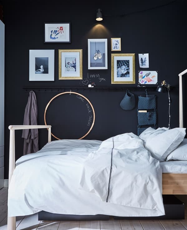 La cama de un adulto contra una pared oscura, donde se muestran dibujos del niño y barras de pared con organizadores colgantes.