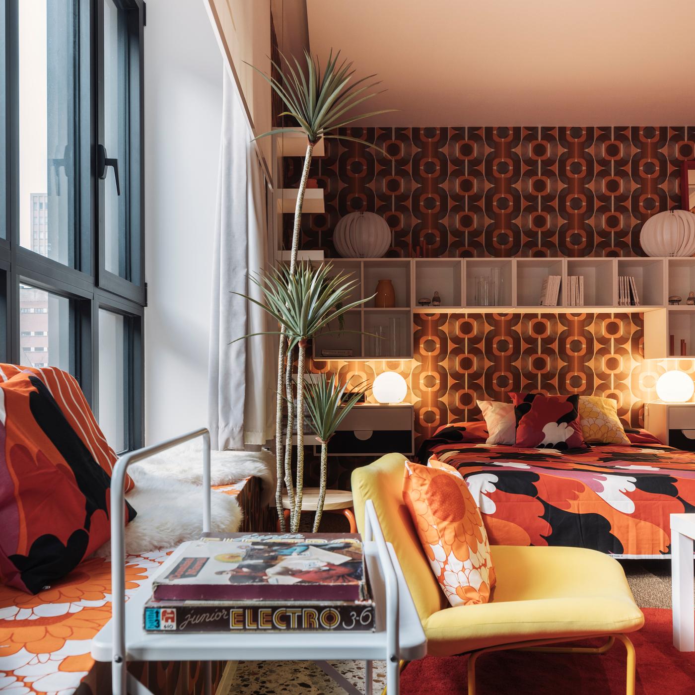La chambre rétro présente une palette de couleurs orange et marron, un motif floral sur la literie, une fenêtre de siège avec des coussins, de grandes plantes, un papier peint rétro, des tables de chevet avec des lampes rondes, des étagères murales avec des livres et des décorations, et une chaise jaune à côté d’une table avec des magazines.