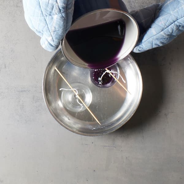 La cire fondue violette est soigneusement versée dans des récipients en verre munis de mèches fixées, comme si un projet de bougies DIY à la IKEA était en cours de réalisation.