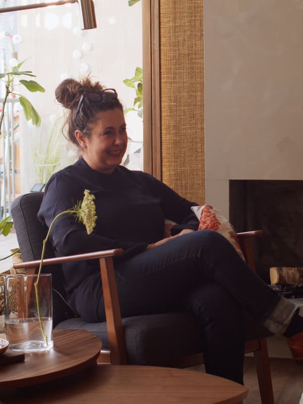 La designer d’intérieur Chiara Effroi Lutteri, assise dans une chaise devant une fenêtre avec des tables et des fleurs dans un vase.
