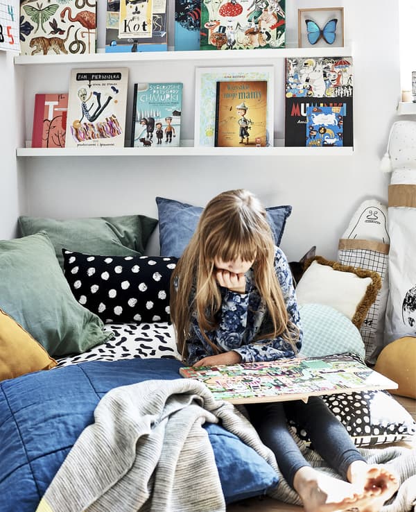 La Hania llegint al seu llit amb teixits colorits i llibres en un prestatge per a quadres.