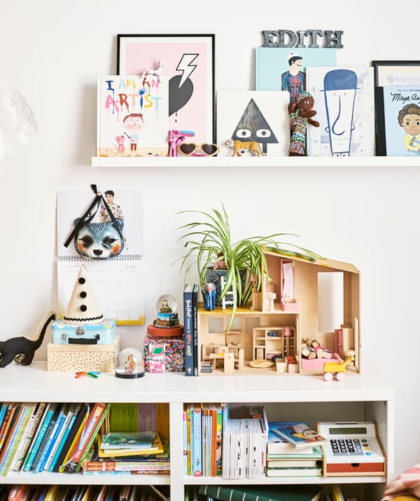 La libreria di una bambina piena di libri e giochi, tra cui una casa delle bambole e delle scatole. Alla parete, una mensola con dei libri.