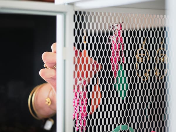 La main d’une personne portant des bracelets et bagues en or, et ouvrant la porte en treillis métallique blanc d’une armoire BAGGEBO.
