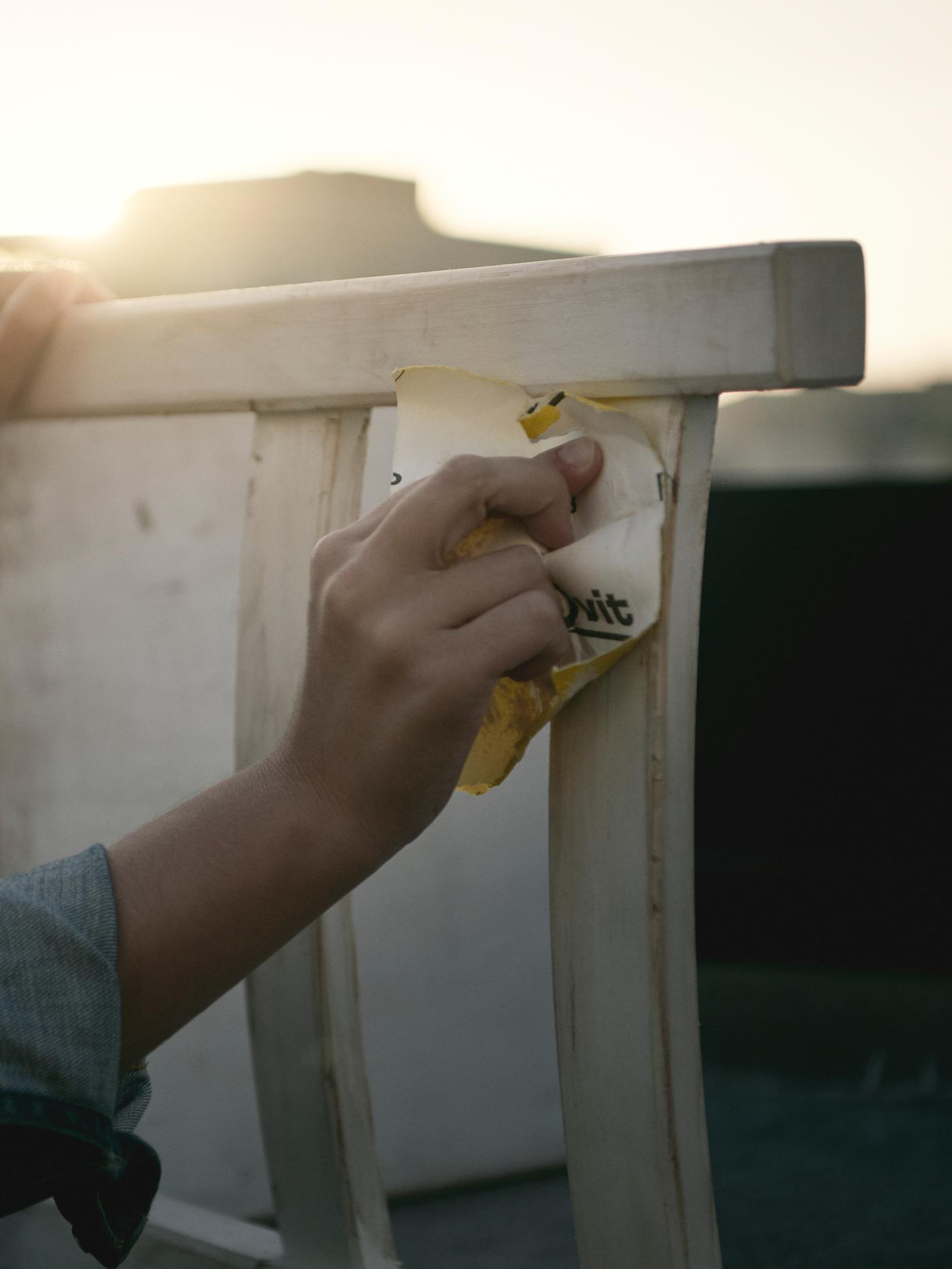 La mano de alguien con las mangas remangadas lijando el respaldo de una silla de madera blanca mientras está al aire libre bajo el sol de la tarde.
