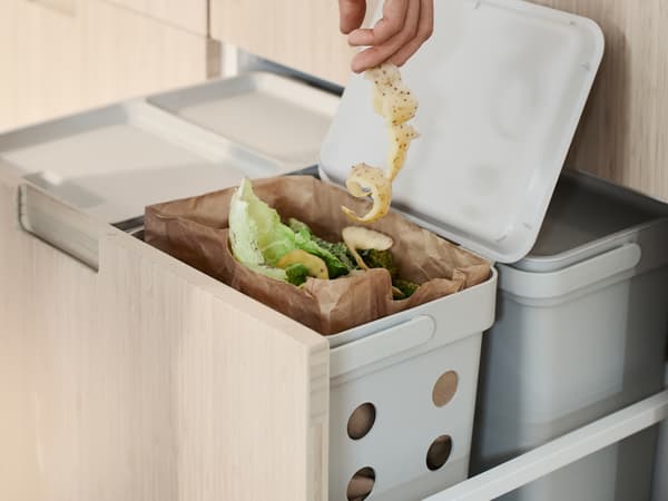 La mano de una persona metiendo una larga ristra de cáscaras de papas en un contenedor HÅLLBAR que contiene residuos orgánicos de cocina.