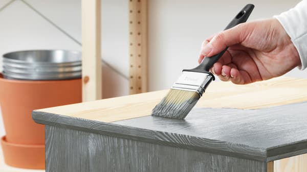 La mano de una persona sostiene un pincel y aplica pintura gris a la superficie superior de un mueble de pino sin tratar.