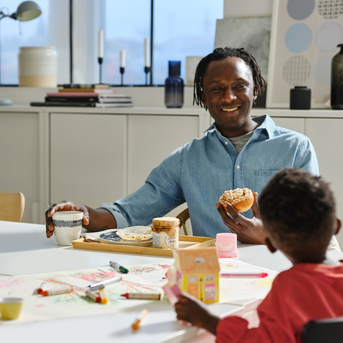 La o masă albă, un adult ține în mână o cană FRIKOSTIG albă/cu model și o chiflă, în timp ce un copil se joacă cu cariocile și hârtia din colecția MÅLA.