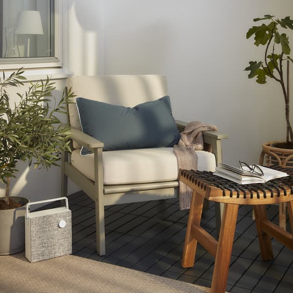 L’accogliente angolo di una terrazza con una poltrona grigia con cuscini bianchi, uno sgabello di legno, una cassa bluetooth bianca e un plaid rosa - IKEA