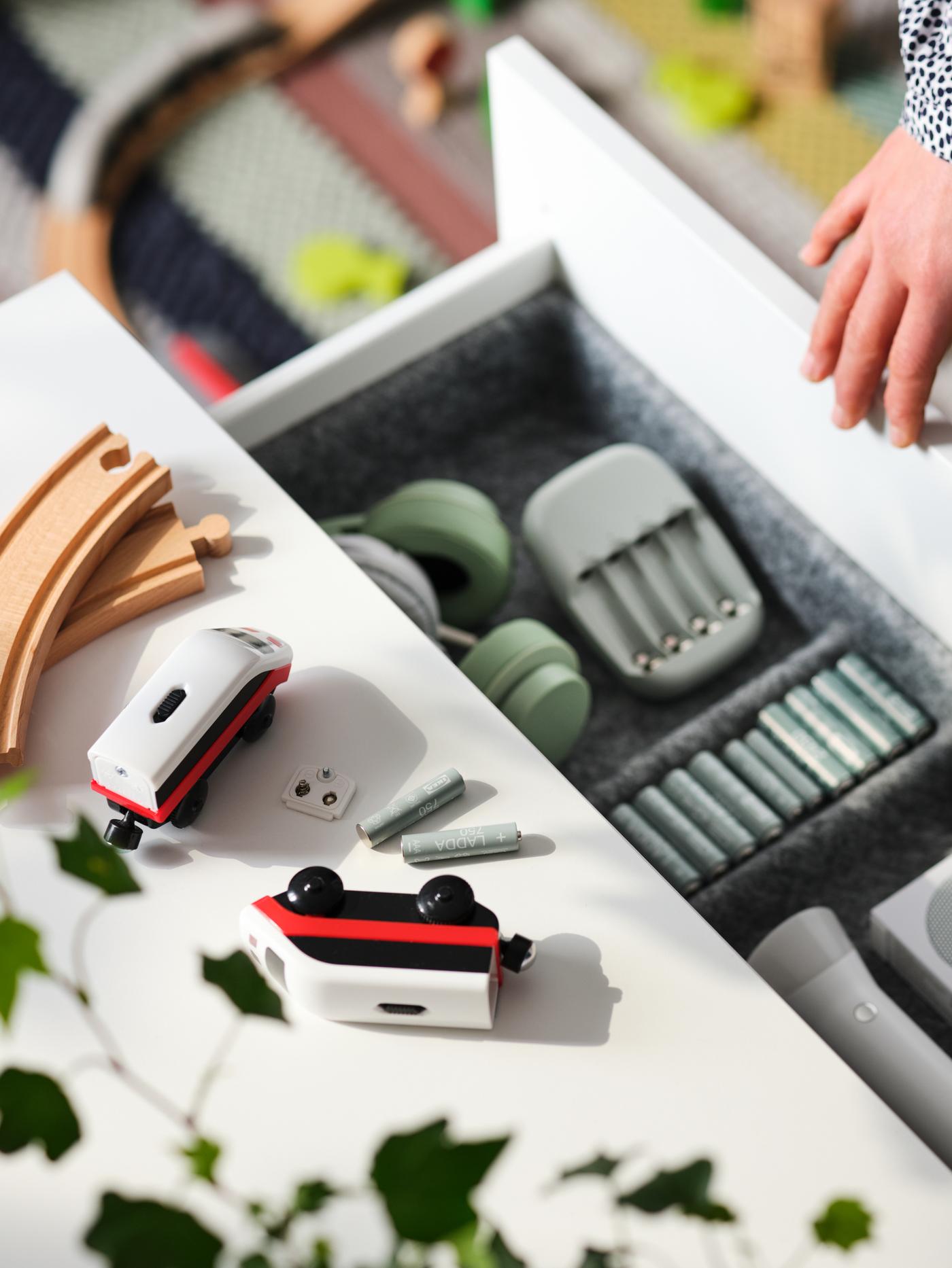 LADDA rechargeable batteries and charger in the open drawer of a white chest of drawers on which lie toy trains and tracks.