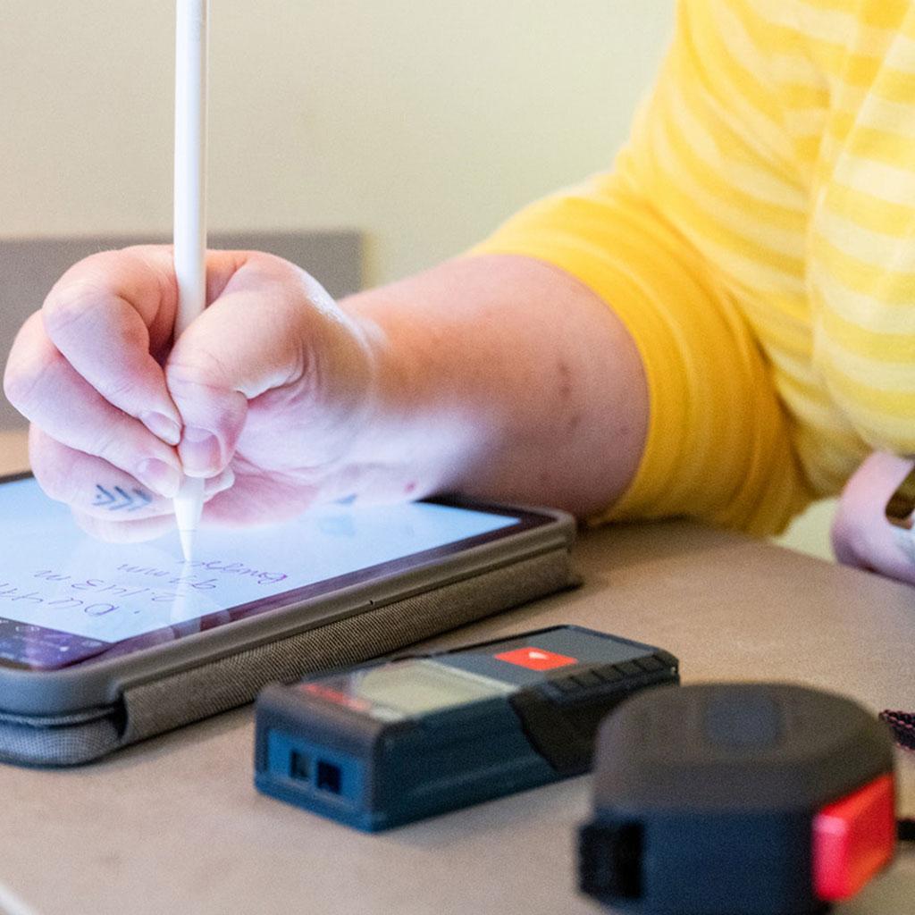 Lähikuva tabletista, lasermitasta ja mittanauhasta. Henkilön kädessä styluskynä tabletin yllä.