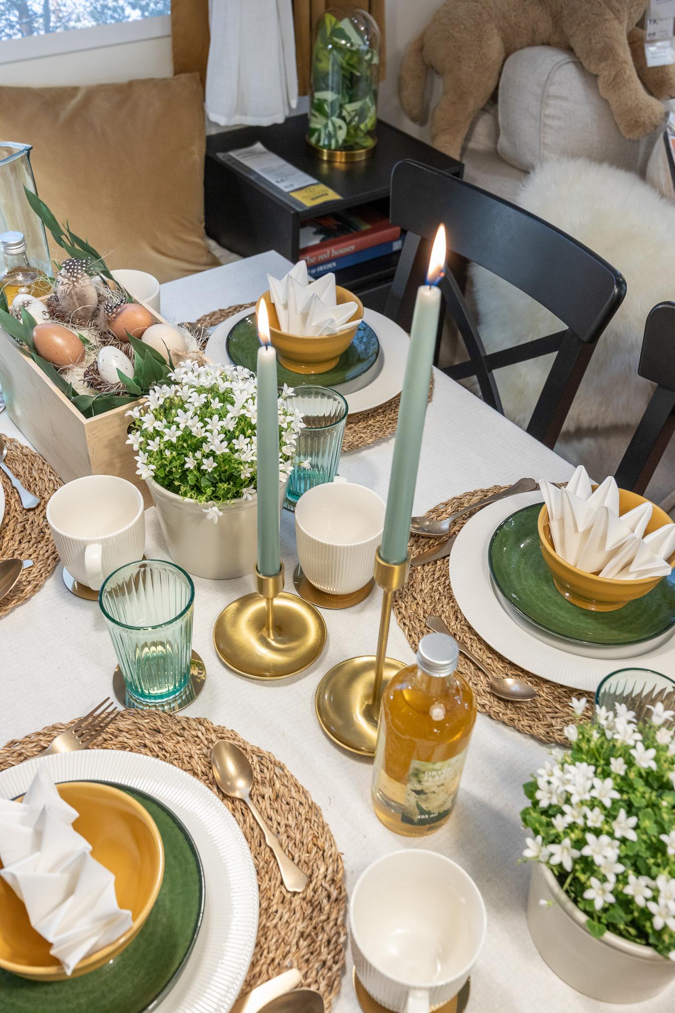 Lähivõte lihavõttelauast, mis on kaetud roheliste ja valgete taldrikute, kollaste kausside, punutud lauamattide, rohekat tooni klaaside ning täpiliste munade ja rohelusega täidetud dekoratiivse puidust karbiga.