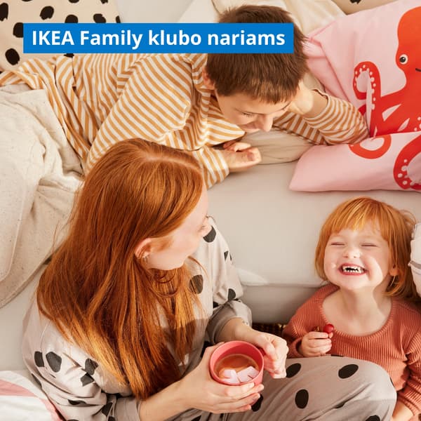 Laiminga šeima svetainėje šalia kampinės sofos: raudonplaukė mama su kakavos puodeliu rankose žiūri į besijuokiančią dukrytę, o vyresnis brolis stebi seserį gulėdamas ant sofos.