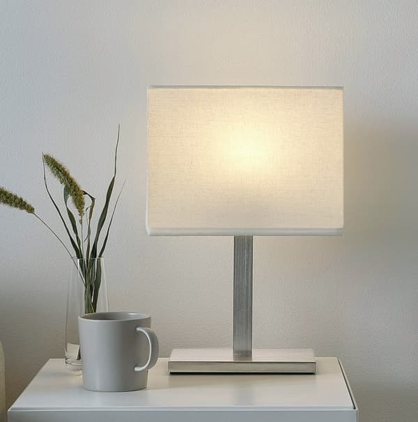Lampa stołowa TOMELILLA z białym kloszem i stalową nogą stojąca na białej szafce nocnej.  