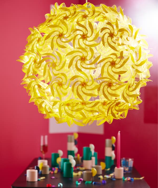 Lampă suspendată galbenă și mare, realizată din spirale inspirate de origami, conectată la o formă rotundă, emițând o lumină difuză într-o încăpere roșie.  