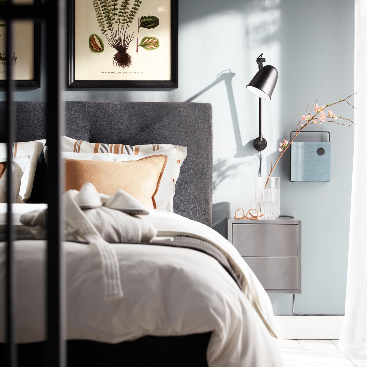 Lampada da parete nera SKURUP, un mobile EKET grigio scuro e una cassa Bluetooth montata a parete accanto a un letto IDANÄS grigio scuro.