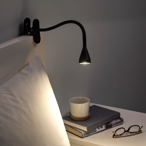 Lámpara de mesa NÄVLINGE negra sobre una cama y apuntando a dos libros y un par de anteojos de lectura.