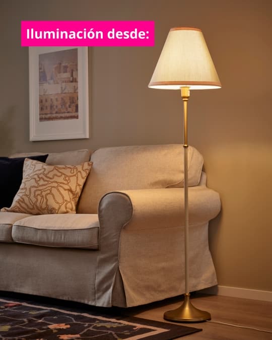 Lámpara de pie con pantalla clara junto a un sofá beige en una sala de estar acogedora. Texto en imagen dice: “Iluminación desde:”.