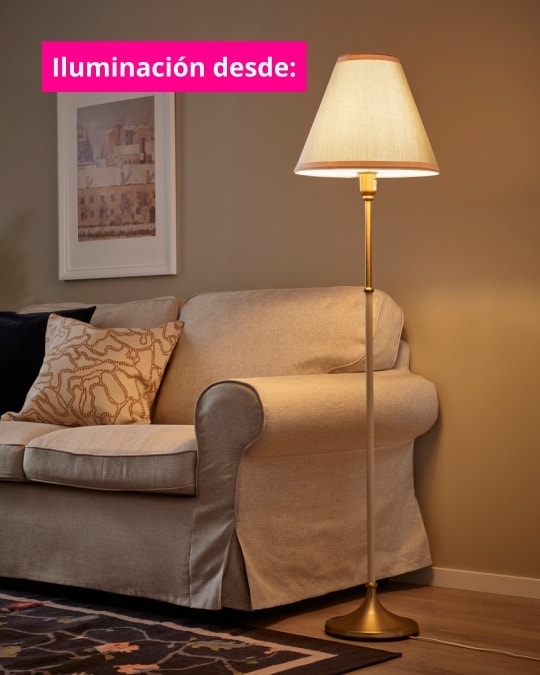 Lámpara de pie con pantalla clara junto a un sofá beige en una sala de estar acogedora. Texto en imagen dice: “Iluminación desde:”.