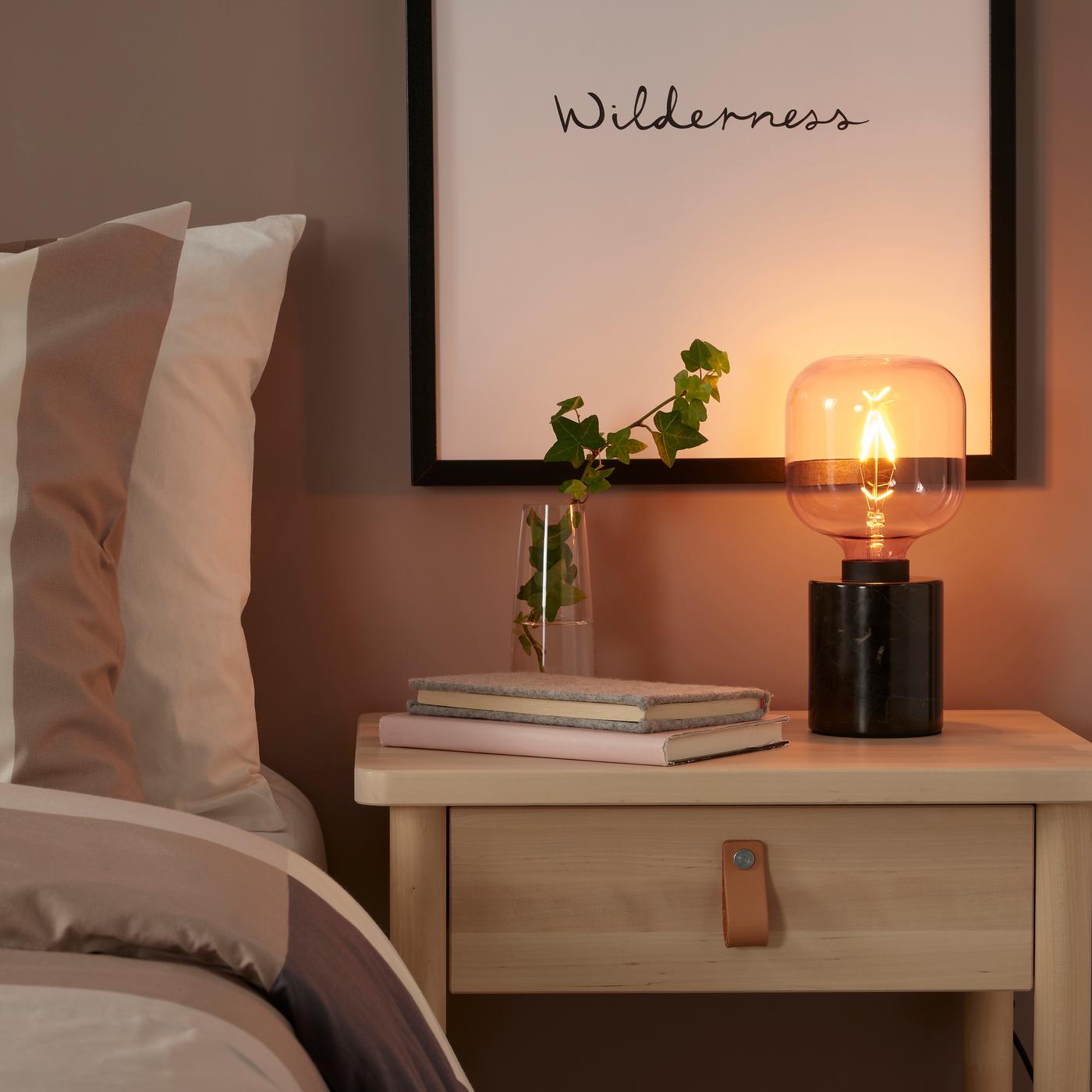 Lámpara LUNNOM con base negra y ampolleta LED. Está sobre una cómoda de madera junto a dos libros y una planta en un macetero de vidrio.