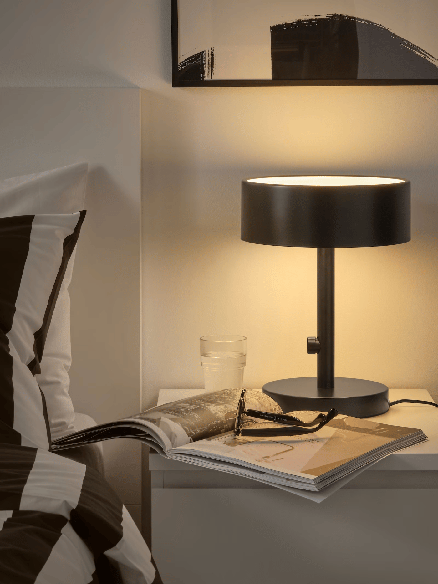 Lampe de chevet noire alluméesur une commode blanche à coté d'un lit...
