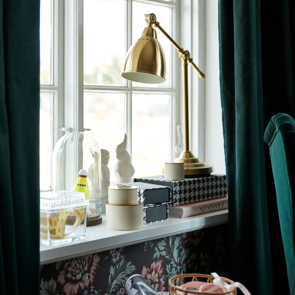 Lampe de travail IKEA BAROMETER couleur laiton près d’une fenêtre avec rideaux SANELA en velours vert foncé.
