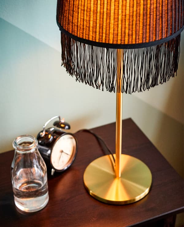 Lampe dorée à franges, réveil et verre d’eau, sur une table de chevet.