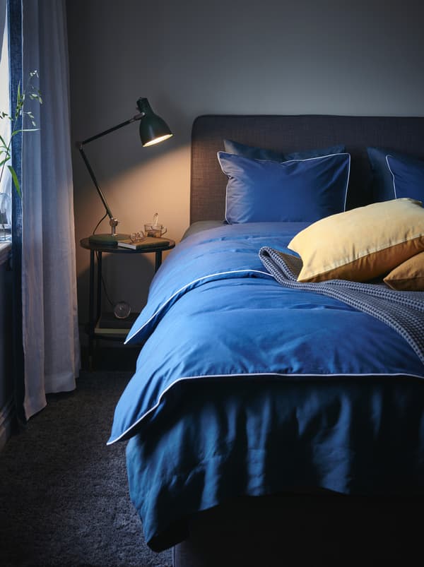 Lampu bacaan yang dinyalakan di atas meja sisi katil. Di sebelahnya terdapat katil dengan duvet berwarna biru, beberapa bantal hiasan dan alas katil berwarna kelabu.