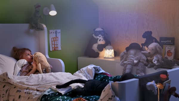 Lampu dinding FUBBLA dan lampu meja SOLSKUR mencerahkan bilik tidur yang nyaman sambil seorang kanak-kanak di katil berbisik kepada mainan patung kegemarannya.