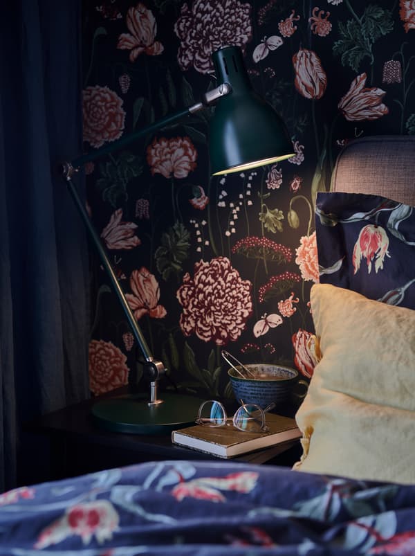 Lampu yang dinyalakan di atas meja sisi katil dengan koleh, cermin mata dan buku. Di sebelahnya terdapat katil dengan duvet berwarna biru dan bantal berwarna kuning.