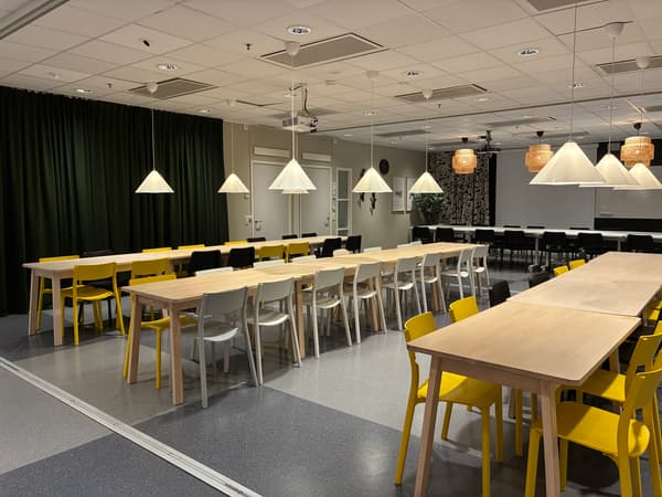Långbord med träskivor och mixade stolar i vitt, svart och gult, upplysta av flera hänglampor i en ljus matsal.