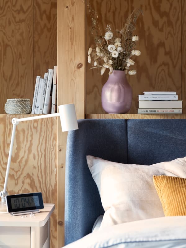 L’angolo arrotondato di un letto TUFJORD blu davanti a una parete rivestita in legno, con libreria integrata e comodino. Sul comodino c'è una lampada da lavoro NYMÅNE. 