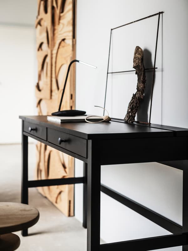 Langs væggen i et hvidt rum står et slankt, sort HEMNES skrivebord og en stor kunstdekoration af ubehandlet træ.