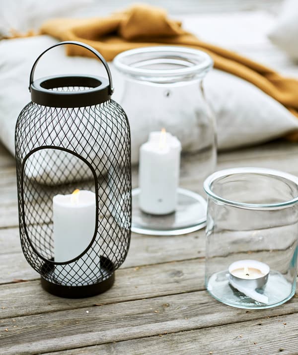 Lanterne en treillis noire avec une bougie bloc à côté de deux bocaux en verre avec des bougies, à côté d’un coussin et d’un plaid jaune posés sur le pont.