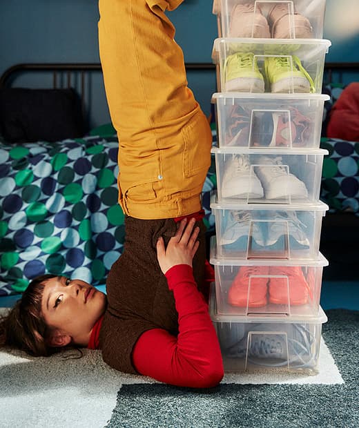 Lány egyensúlyoz a hátán, mögötte SAMLA dobozok, színes cipőkkel.