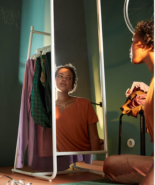 Lány ül a padlón, tükörben nézi magát, mellette ruhák egy ülőkén.