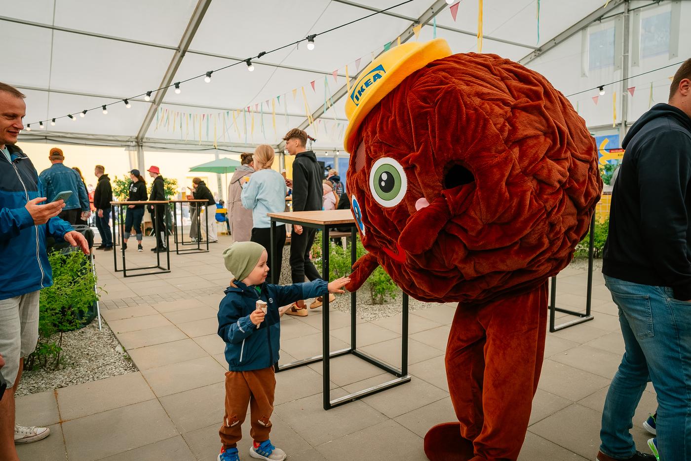Laps sirutab käe lihapallimaskoti poole, kellel on peas kollane IKEA müts, kaunistatud peotelgis, kus taustal on näha külastajaid, laudu ja valguskette.
