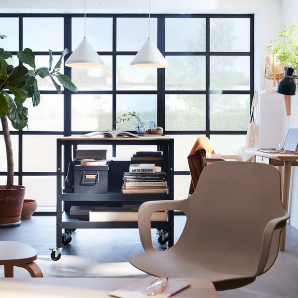 Large black-framed windows with two white pendant lamps, a beige swivel chair and a black/pine trolley on castors in front.