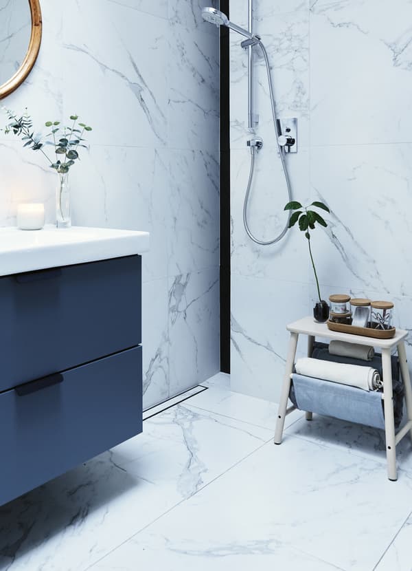 Lavabo gris dans une salle de bain carrelée de marbre blanc au mur, douche murale et tabouret en bois sur lequel sont posés des flacons.