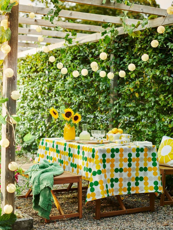 Lavice a stôl NÄMMARÖ prikrytý obrusom BRÖGGAN stoja v záhrade pod pergolou so svetelnou reťazou SOLVINDEN.