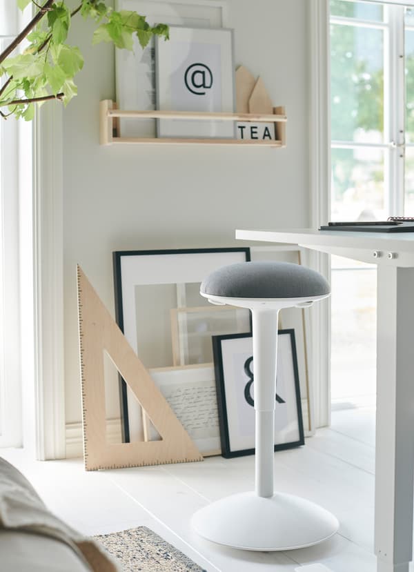 Le bureau assis/debout IKEA SKARSTA blanc possède une manivelle pour régler la hauteur du plan de travail. Le tabouret NILSERIK à coussin gris clair est également réglable pour une assise flexible.
