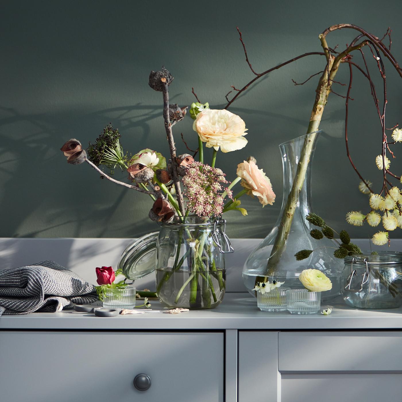 Le dessus d’un buffet gris HAUGA avec diverses fleurs dans des bocaux en verre, des photophores et une carafe en verre transparent STORSINT.