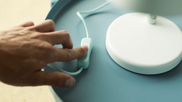 Le doigt d’une personne appuie sur l’interrupteur marche/arrêt du cordon d’une lampe de travail à DEL FUBBLA blanche ; cette dernière est posée sur une table-plateau bleu clair.
