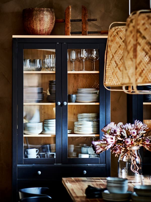 Le long d’un mur, près d’une table à manger, une vitrine HEMNES de couleur foncée à portes en verre est remplie de vaisselle.