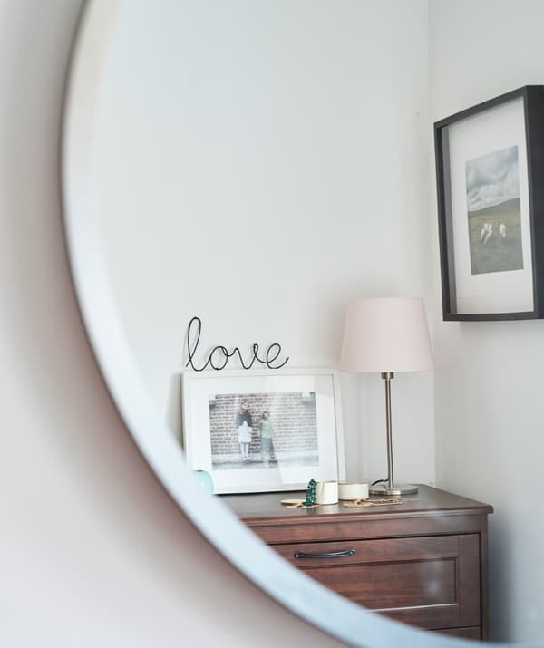 Le reflet d’une commode sur laquelle est exposé un cadre photo, à côté d’une lampe de table avec pied en laiton et abat-jour rose et de petites boîtes à bijoux.