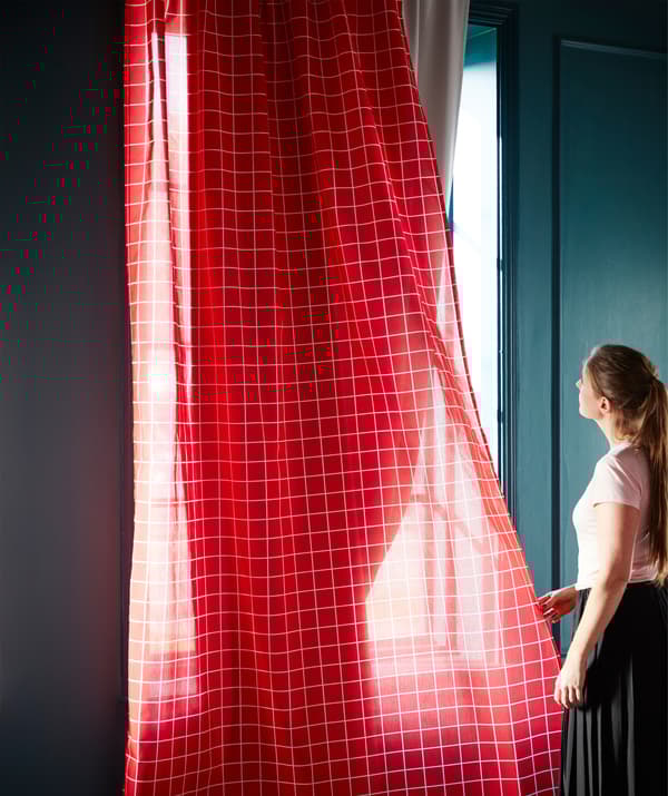 Le tende oscuranti grigie MAJGULL di IKEA bloccano la luce, mentre delle leggere tende rosse la lasciano filtrare. Una donna ha in mano le tende rosse e guarda fuori dalla finestra.