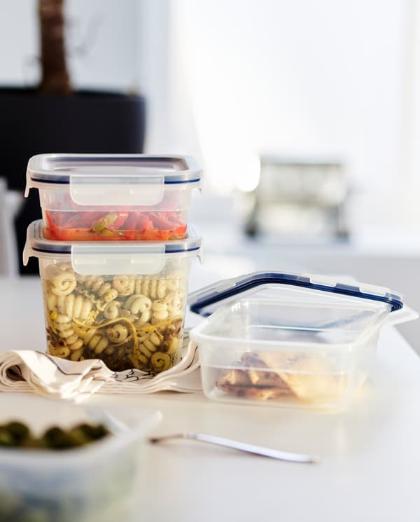 Lebihan makanan di dalam tiga bekas makanan berpenutup plastik, tuala pengesat berwarna hitam/putih dan garpu di permukaan atas kerja.