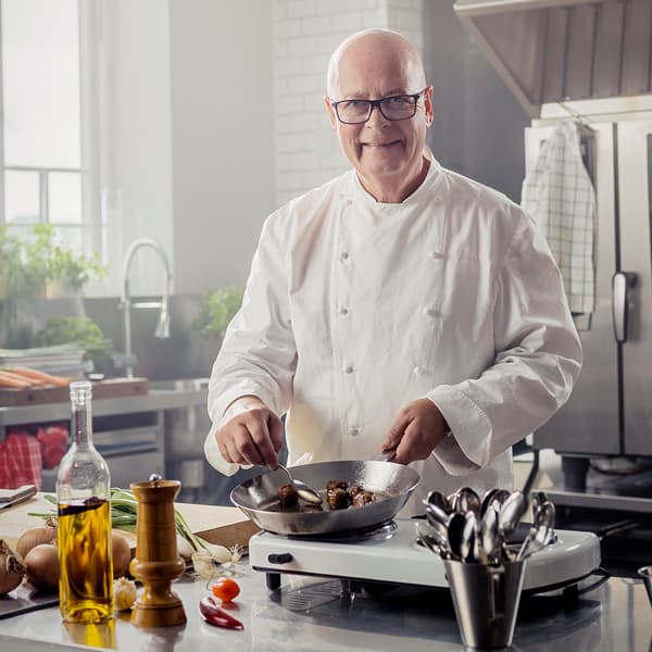 Leende person iklädd vit kockrock som står och steker köttbullar i ett reataurangkök. 