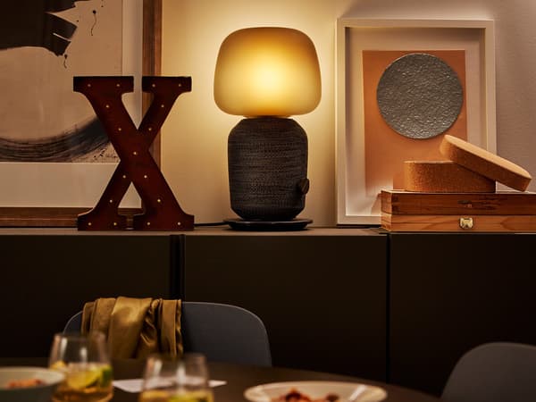 L'enceinte-lampe Wi-Fi SYMFONISK sur une étagère entourée de nombreuses décorations et une table surmontée de verres à vin en avant-plan.