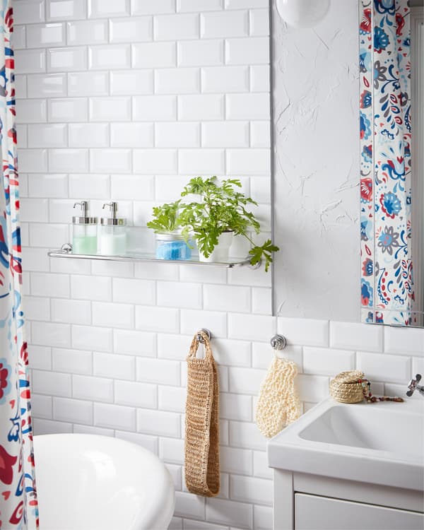 L’étagère en verre VOXNAN et sa bordure en acier chromé, le distributeur à savon et la plante enjolivent l’espace à proximité de la baignoire et du meuble-lavabo.