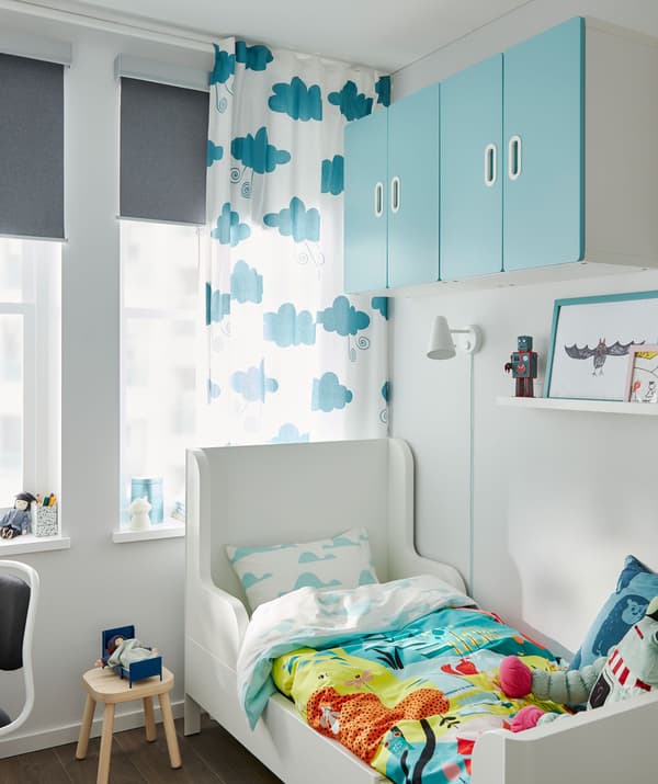 Letto allungabile nell’angolo di una cameretta, copripiumino colorato, pensili alle pareti e una finestra con tende e tende a rullo.